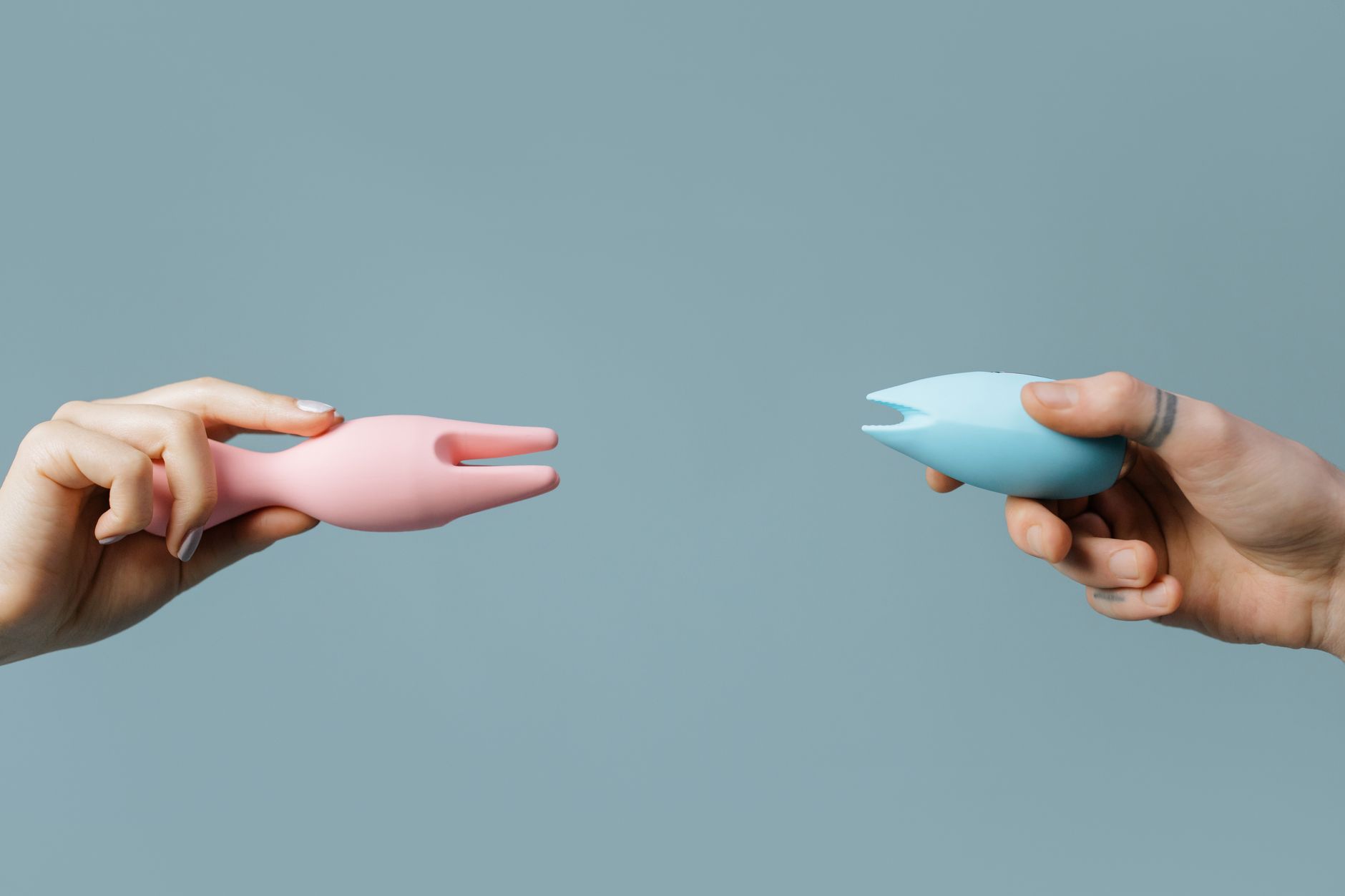 Two hands holding lemon and blue silicone vibrators against a soft pastel background.