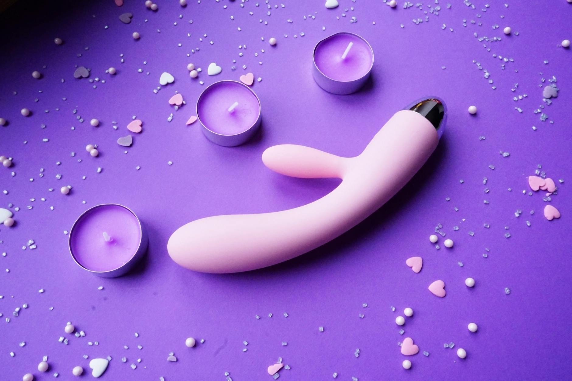 A vibrant pink vibrator on a purple background with romantic candles and heart confetti.